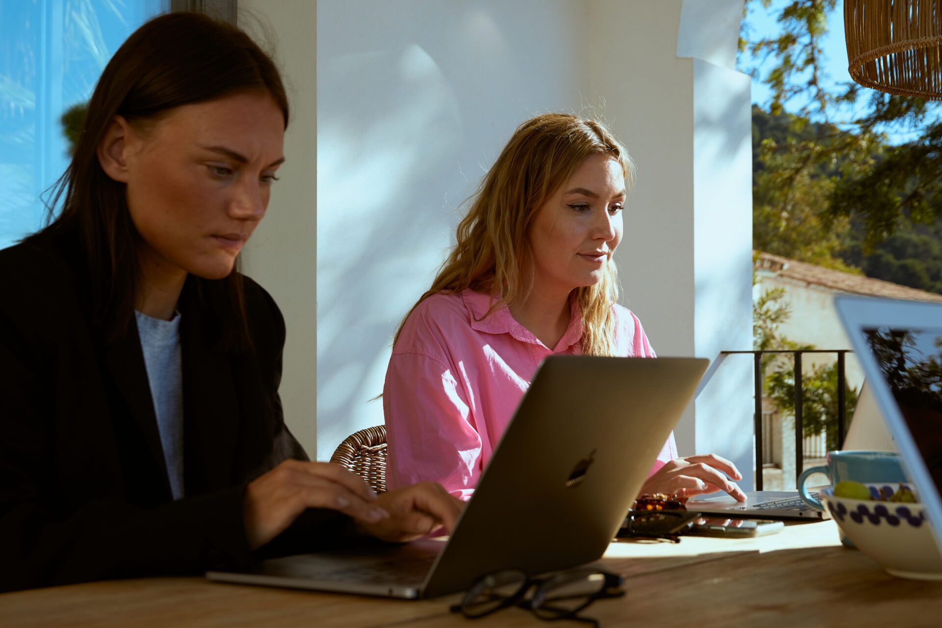 Martina Myrén och Alexandra Åhre sitter bredvid varandra med varsin laptop framför sig som dom arbetar på. De sitter vid ett konferensbord på en veranda och i bakgrunden ser man en blå himmel och gröna träd. Alexandra har på sig en rosa skjorta, medan Martina har en svart kavaj med vit tröja därunder.