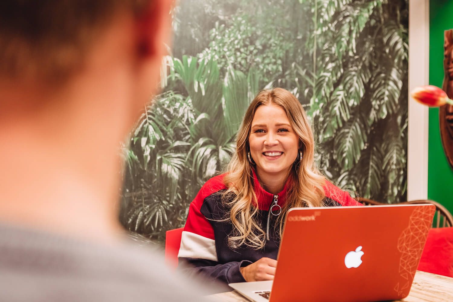 Daniella Ravan sitter leende vid ett bord med en laptop framför sig, med en suddig person i förgrunden. Bakom Daniella ser man en tapet med gröna blad.