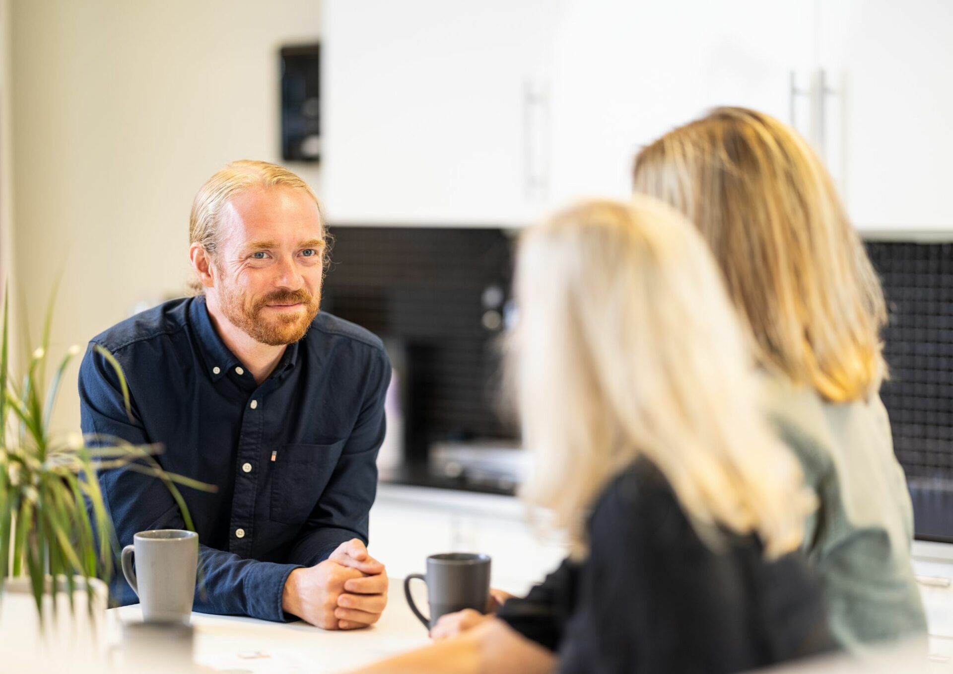 I förgrunden ryggen av två personer med halvlångt blont hår. I fokus en person med mörkblå skjorta, blåa ögon och tillbakakammat blont hår.