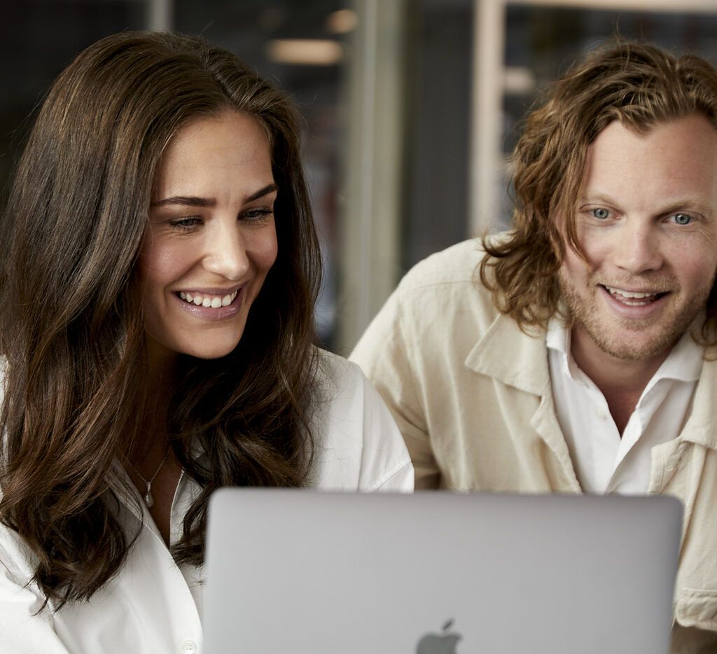 Två leende kollegor samarbetar vid en datorskärm i en varm och modern arbetsmiljö.